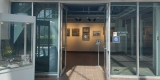 Front of art gallery showing open doors, artwork on the walls and a sign indicating what is happening in the exhibition space.
